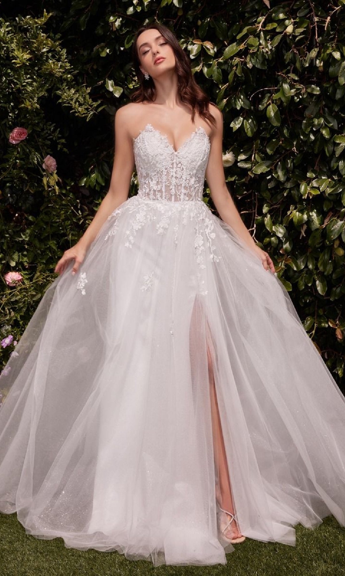 A woman stands outdoors wearing the Strapless Long White Wedding Gown: Ladivine C148W, featuring an off-white lace corset bodice, strapless neckline, tulle skirt with thigh-high slit, and delicate floral appliqués.